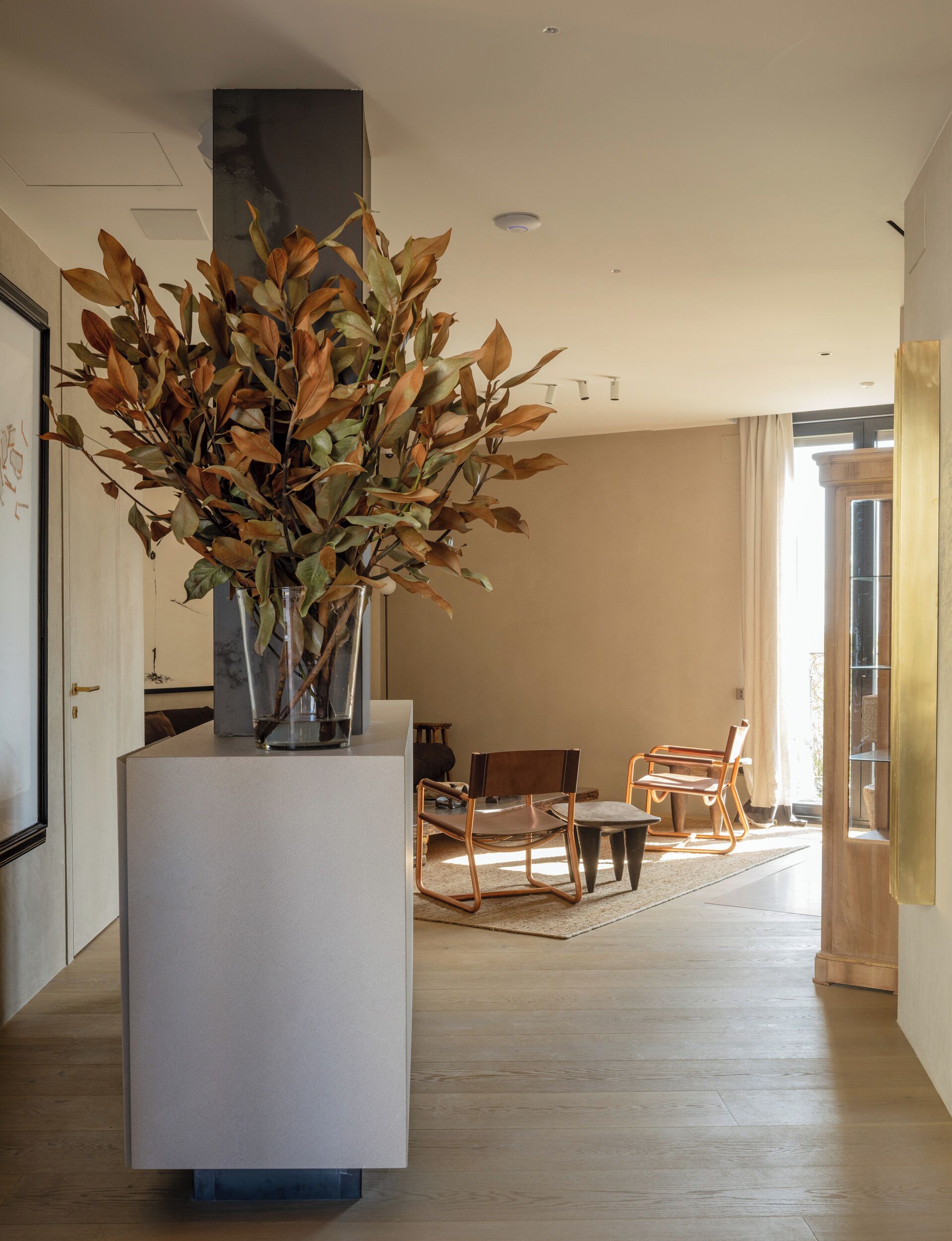 Un salon moderne avec un grand vase rempli de feuilles séchées, posé sur un piédestal, une table à manger en bois et des chaises.