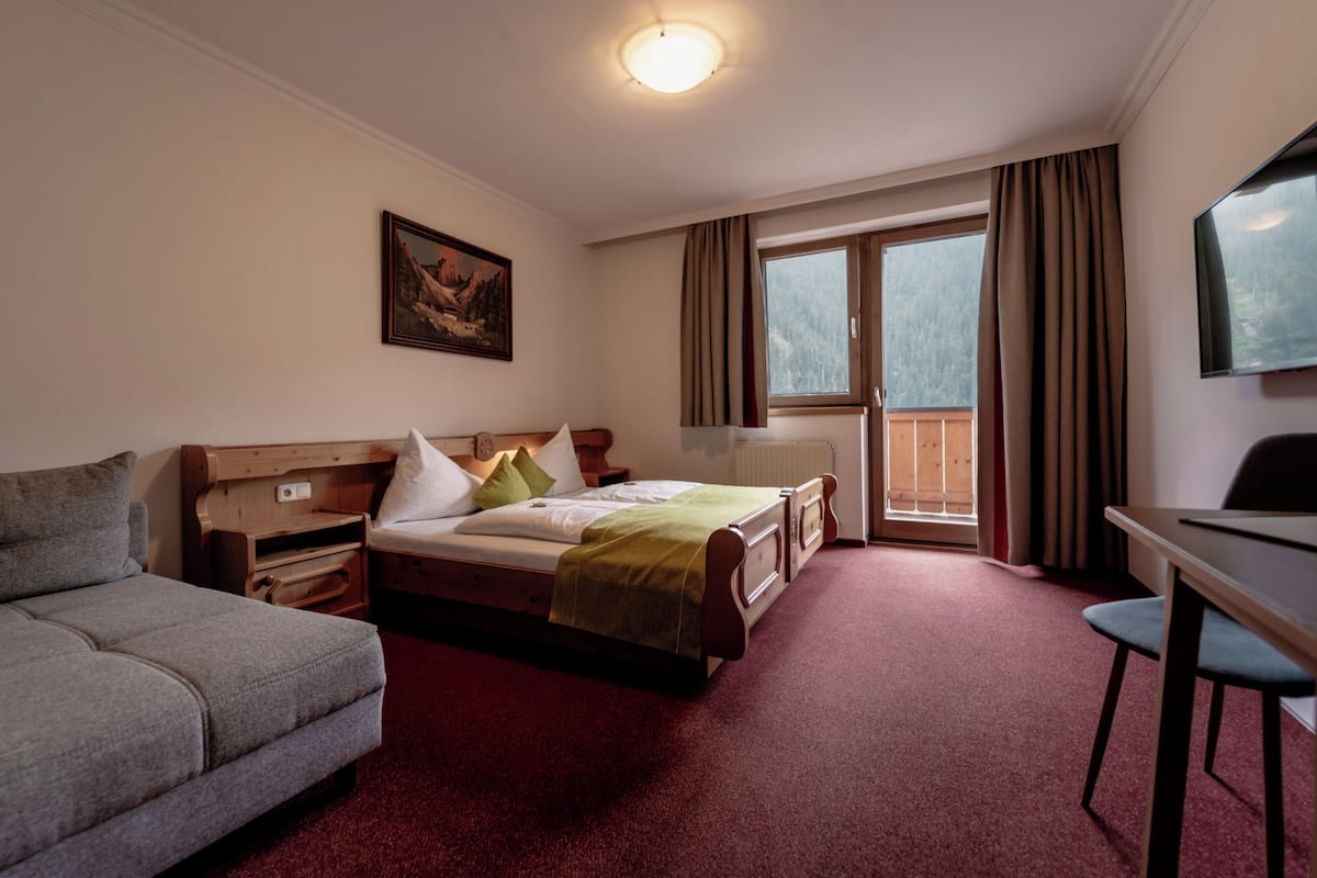 A hotel room with a bed, a sofa, a desk, a window with curtains, a painting on the wall, and a radiator.