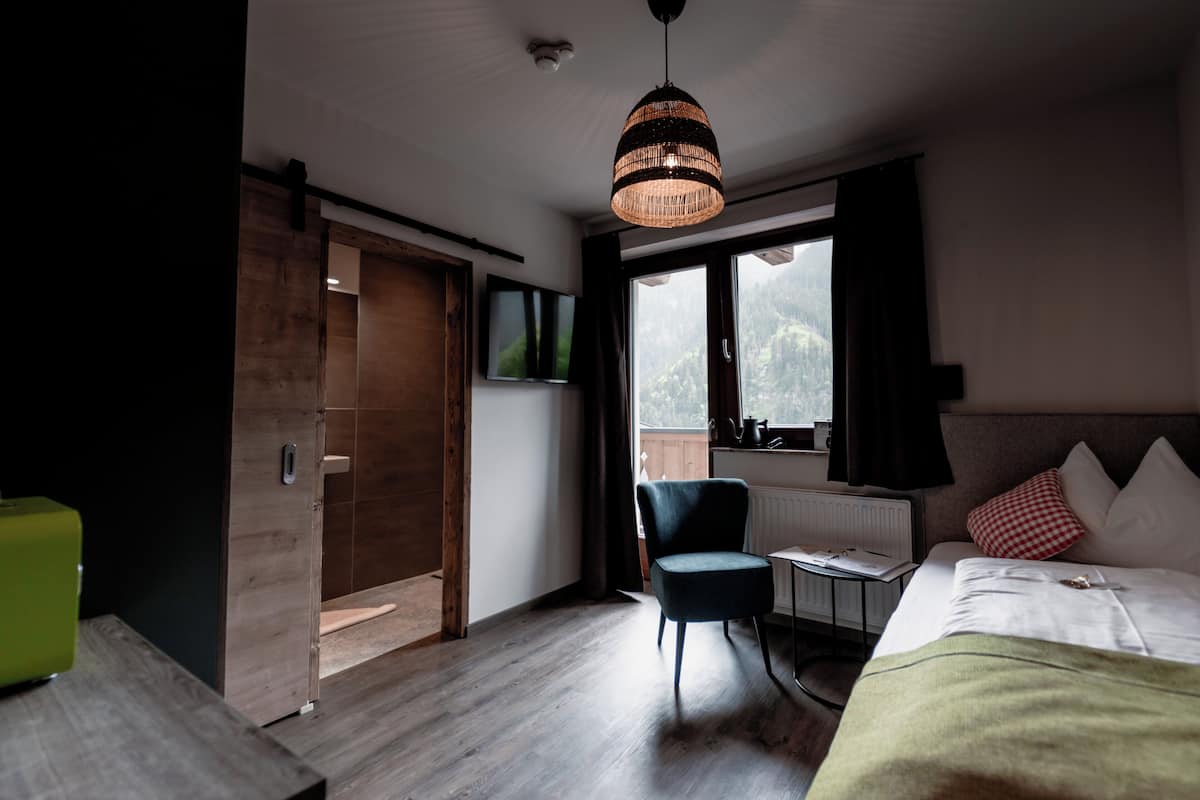 A bedroom with a bed, a chair, a table, a window with a view, and a hanging light fixture.