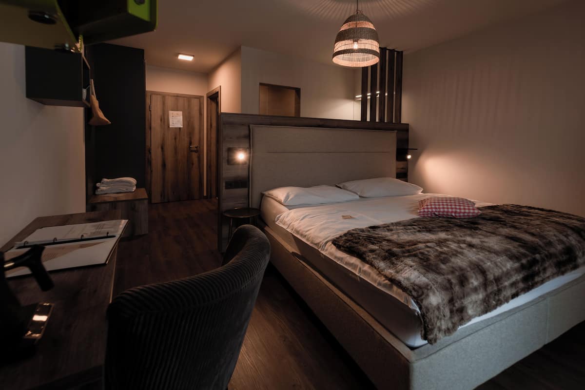 A modern hotel room with a large bed, a desk, a chair, a wardrobe, and a hanging light fixture.