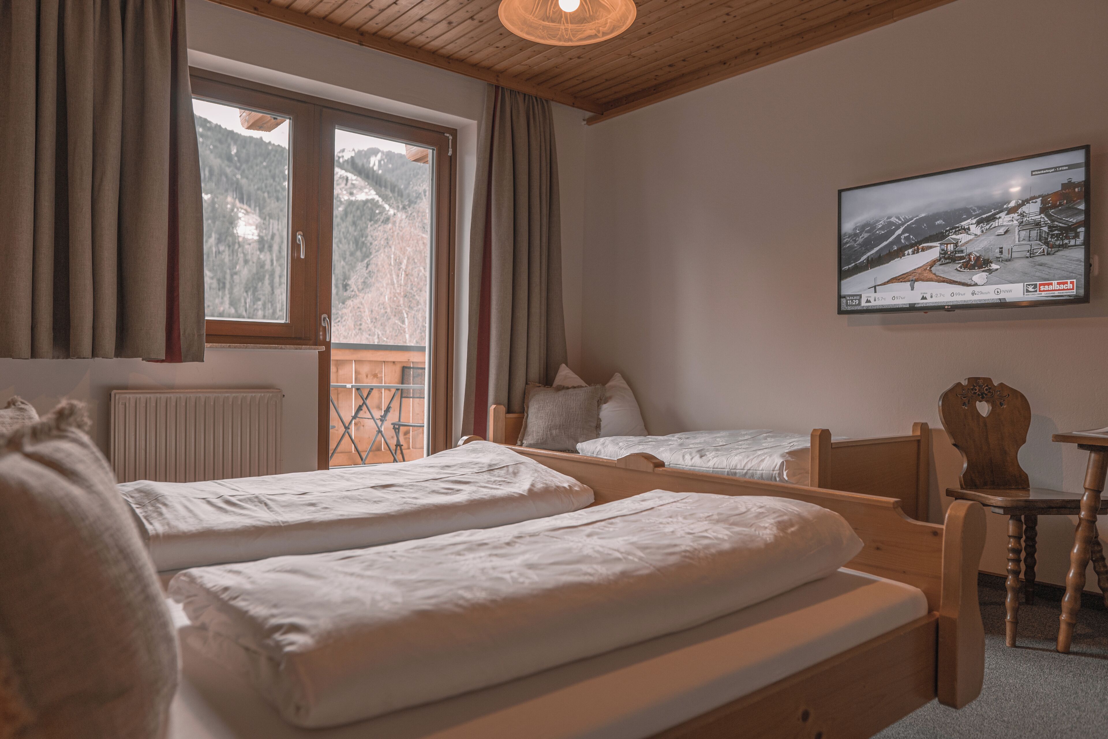 Une pièce comprenant deux lits en bois, une chaise en bois, une table, une fenêtre avec des rideaux et un téléviseur fixé au mur.