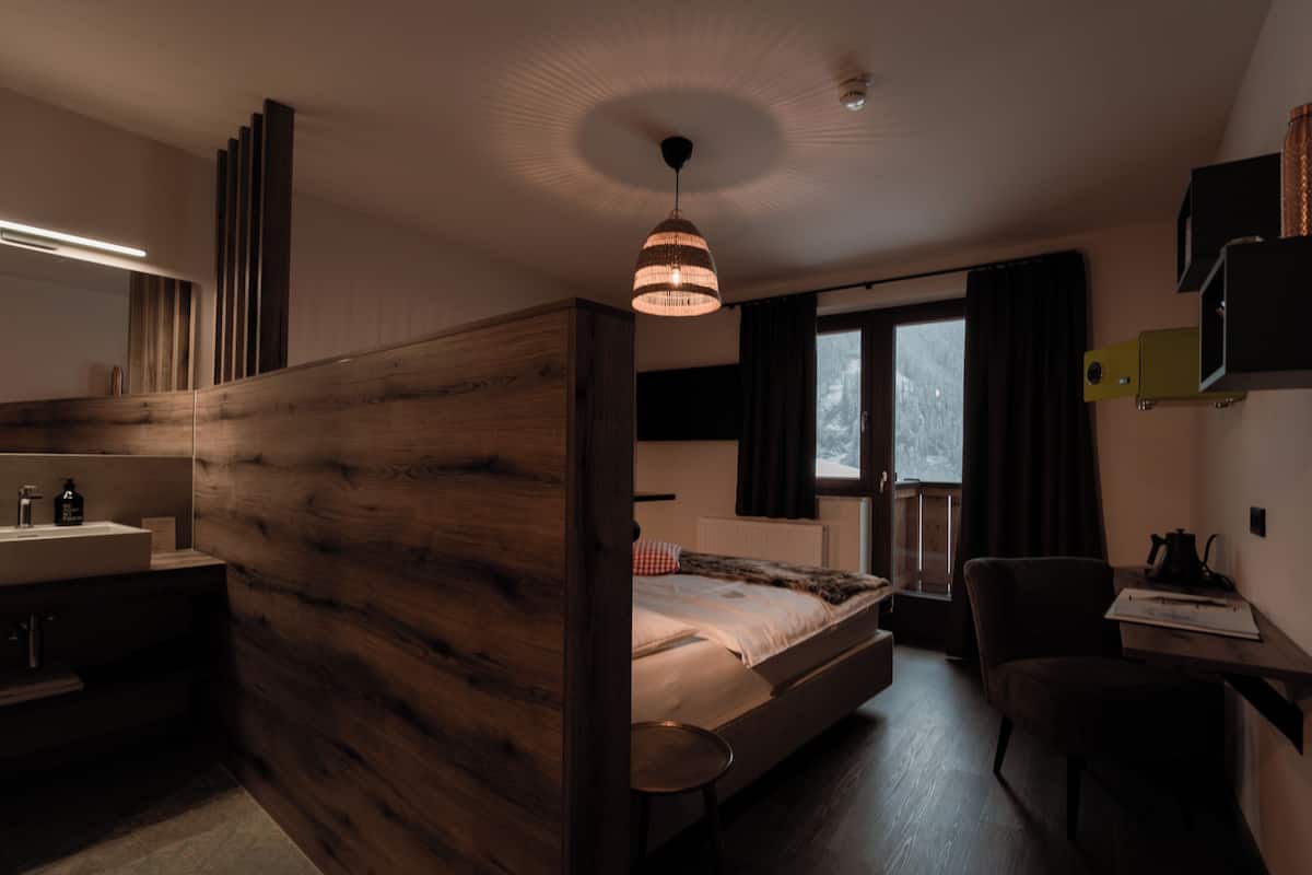 A bedroom with a wooden headboard, a bedside table, a chair, a window with curtains, and a desk with a lamp.