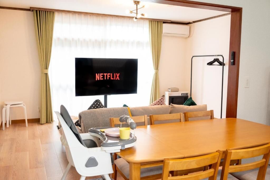 A cozy living room with a wooden dining table, a TV displaying 'NETFLIX', and a sofa with patterned cushions.