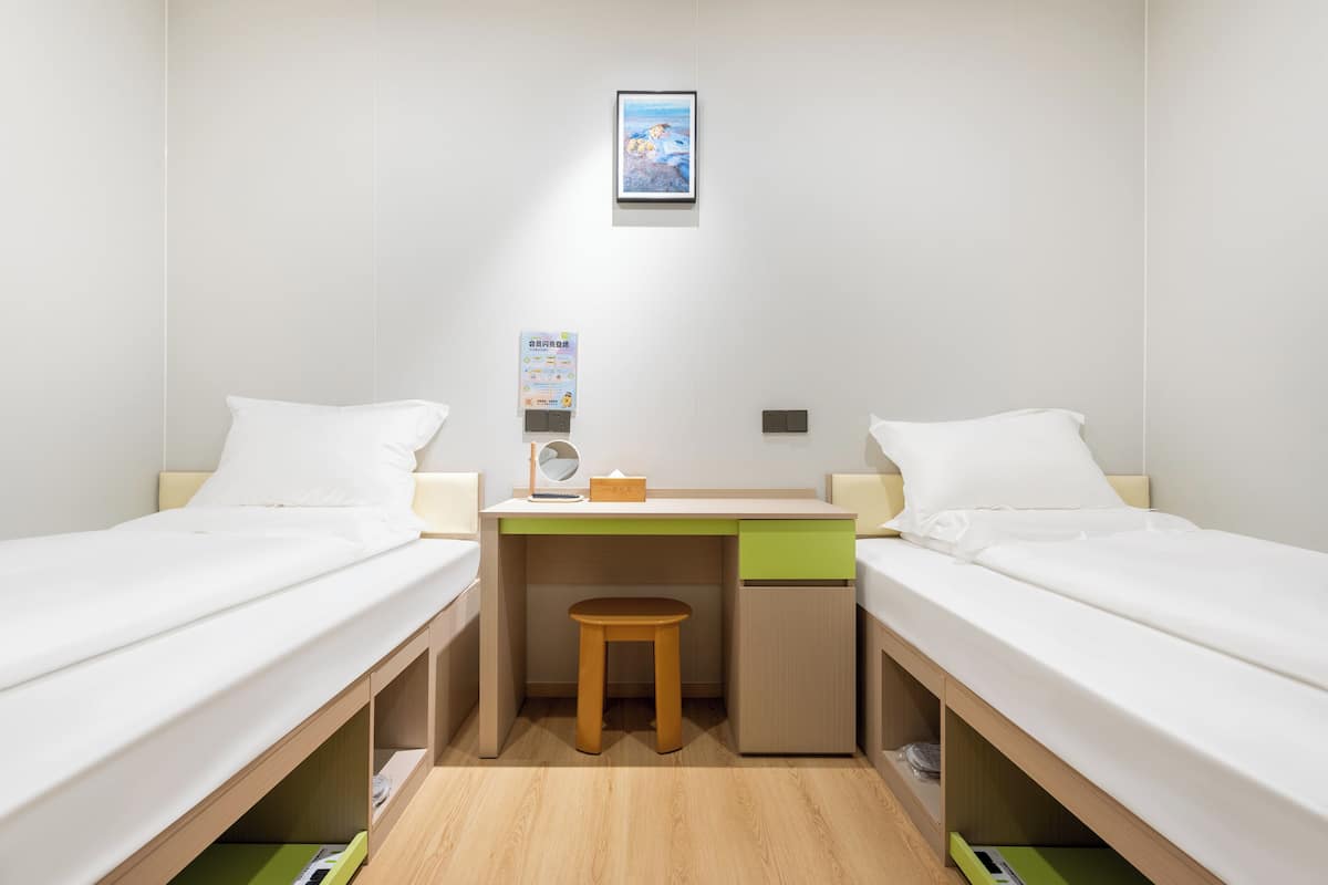 A small, well-lit room with two single beds, a desk with a stool, and a small cabinet.