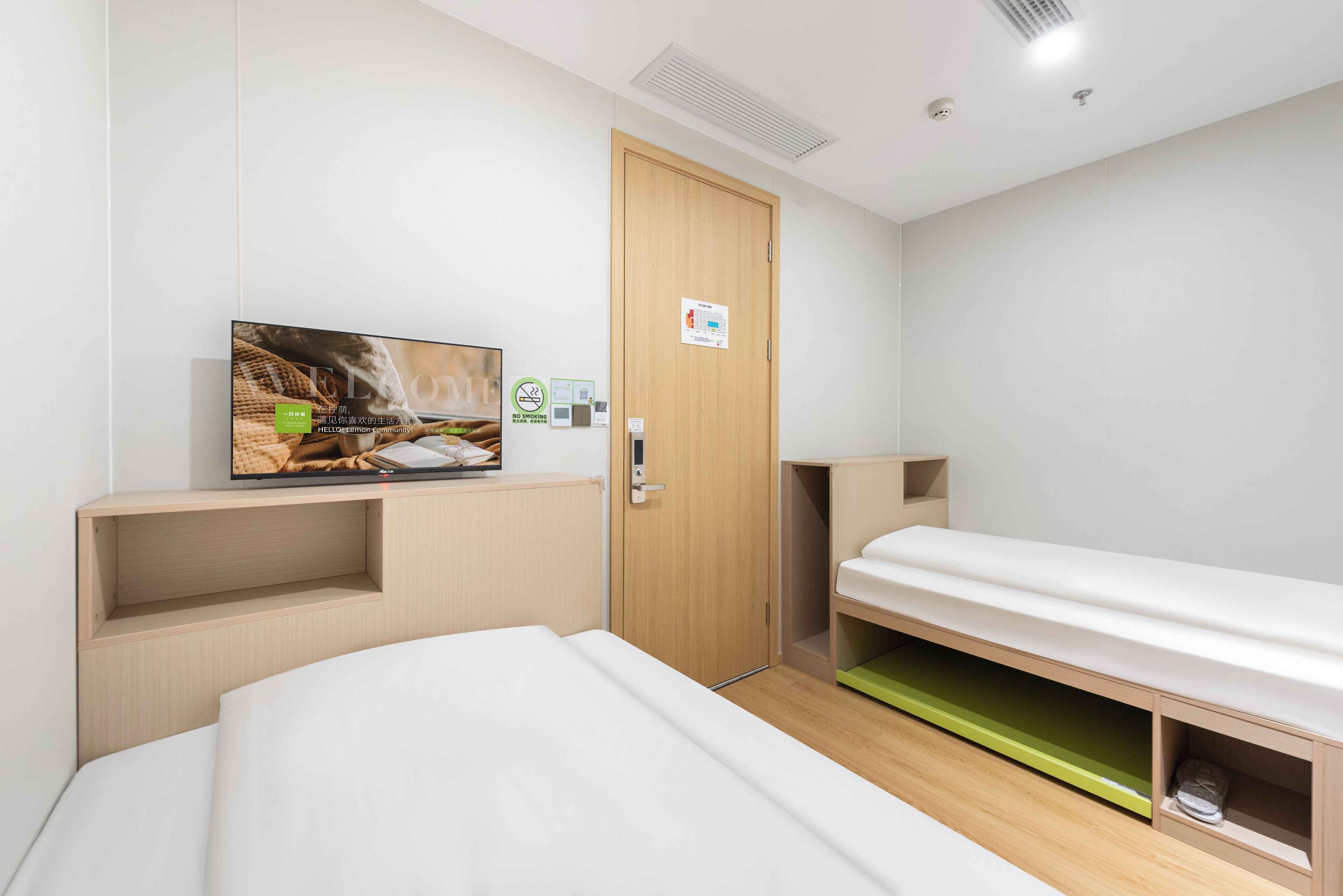 A modern hotel room with a bed, a TV mounted on the wall, a wooden door, and a small shelf.