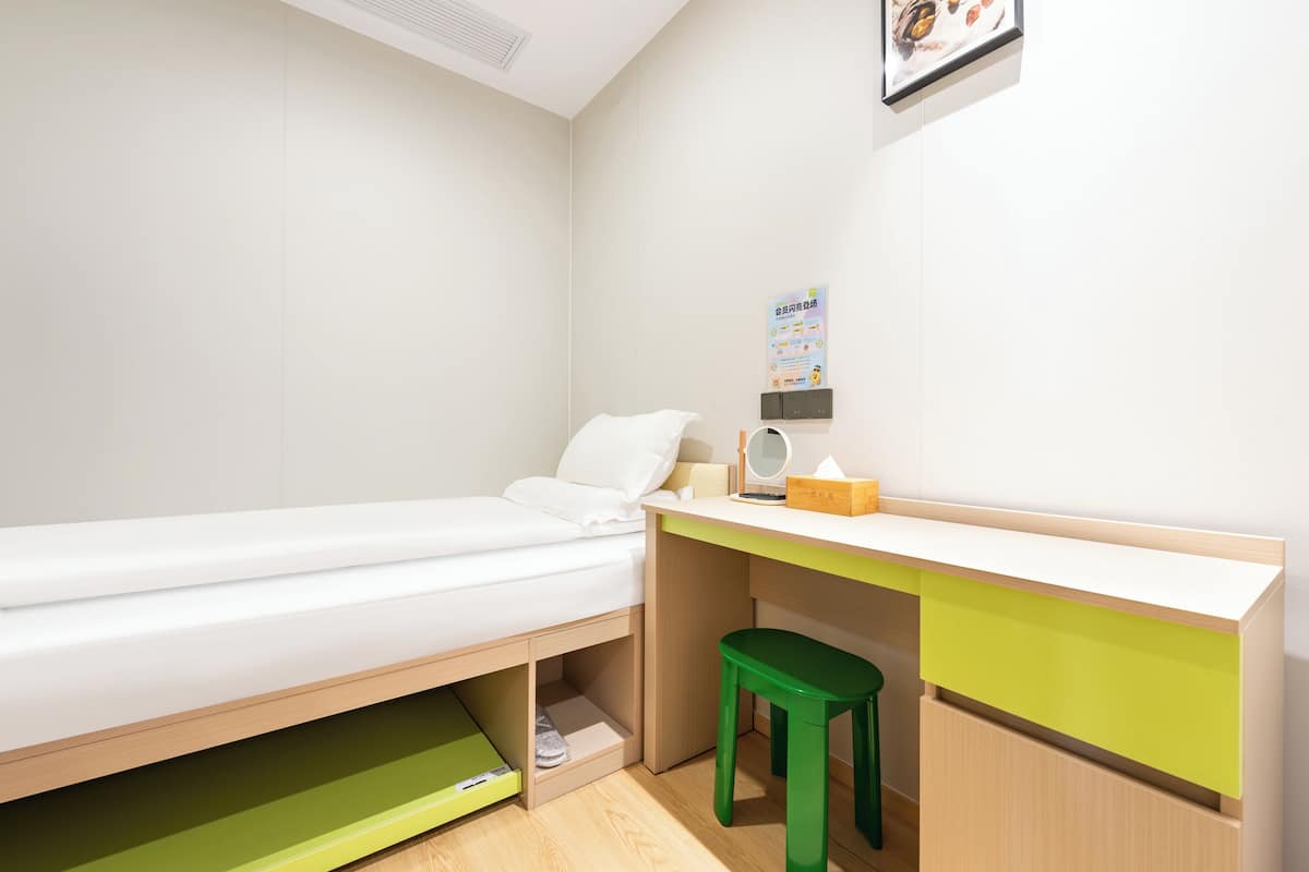 A small, neatly arranged room with a single bed, a desk with a chair, and a small shelf.