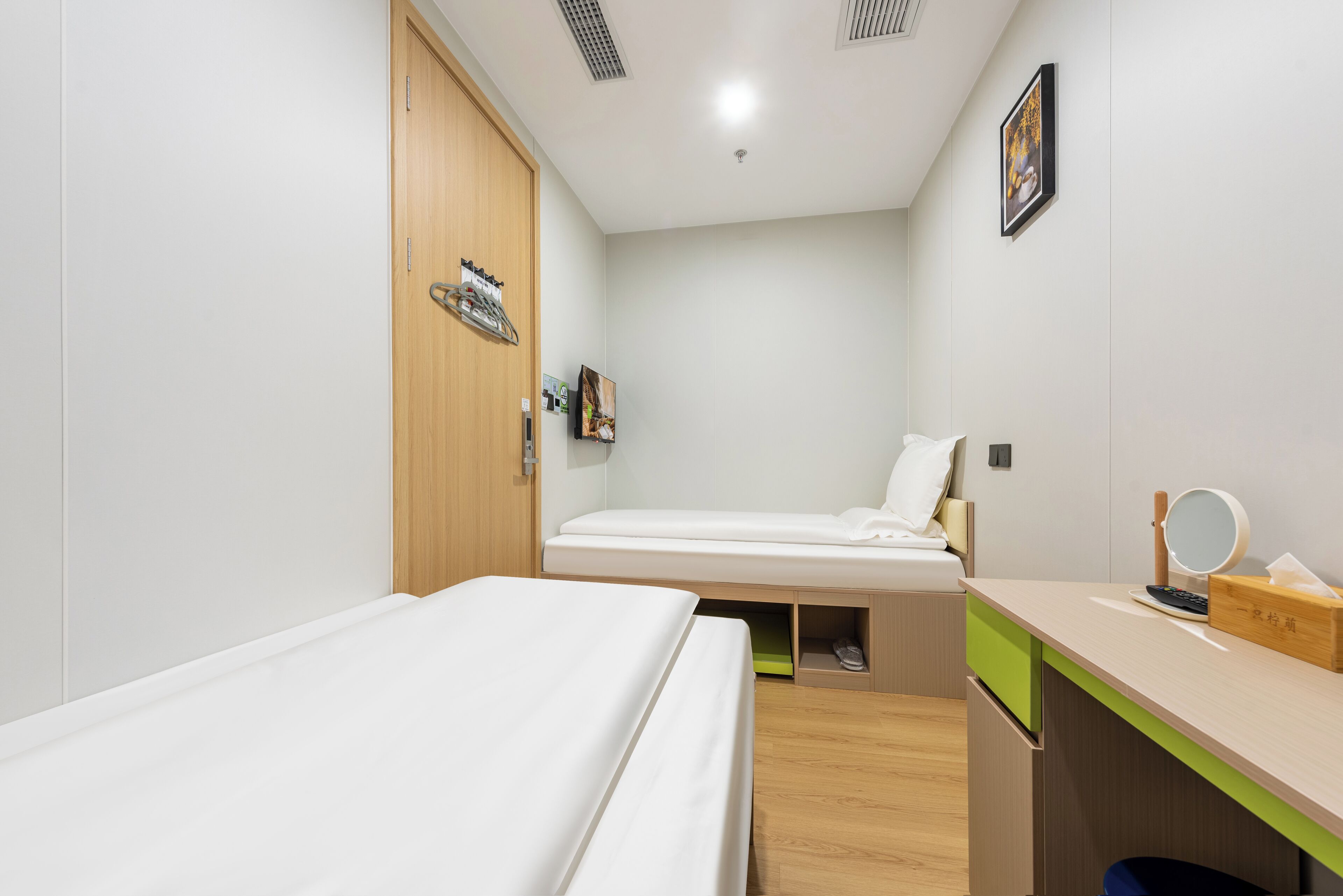 Habitación de hotel pequeña con dos camas individuales, un armario de madera, un escritorio con espejo y un estante con artículos de aseo.