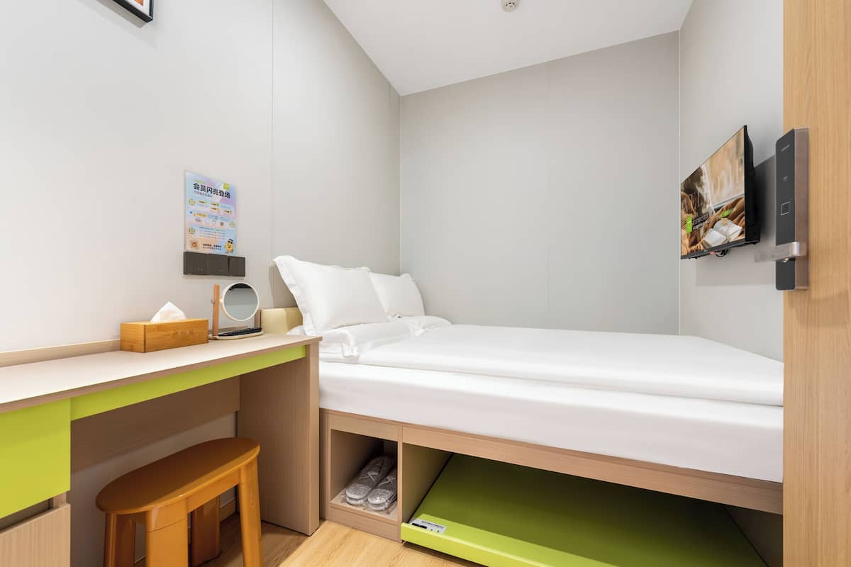 A compact room with a bed, a desk, and a chair. There is a small stool under the desk and a shelf with shoes.