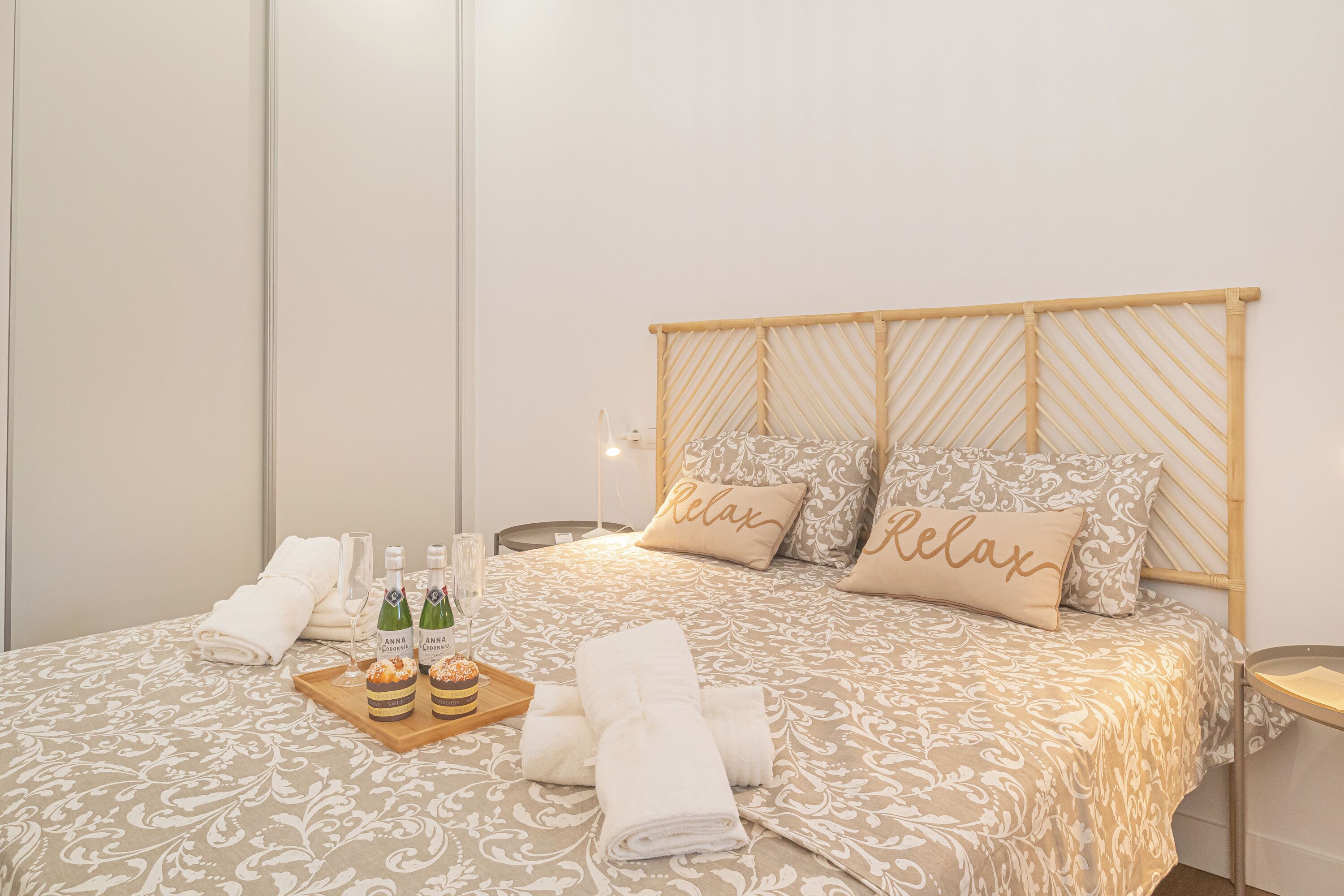 A neatly made bed with a patterned bedspread, two pillows with the word "Relax," a tray with bottles and glasses, and folded towels.