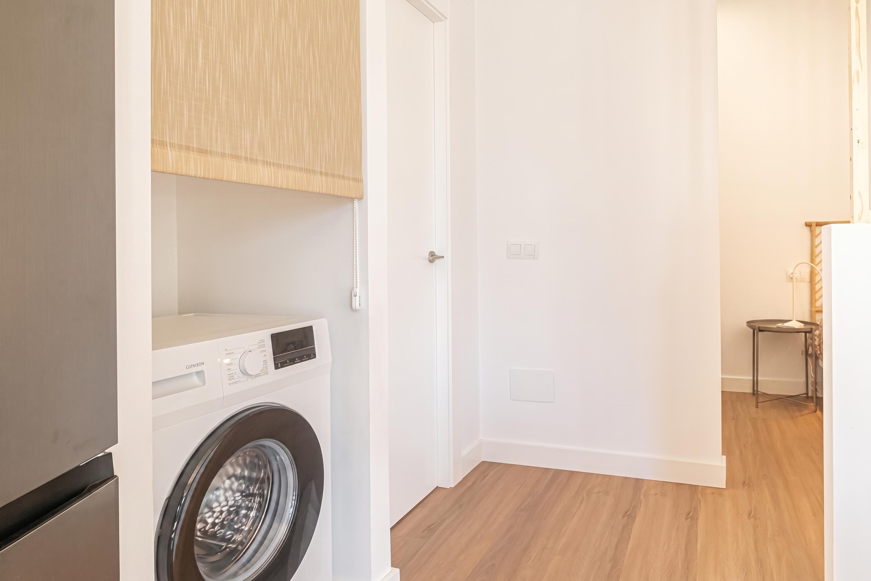 A laundry room with a front-loading washing machine, a wooden cabinet, a door, and a small table with a chair.