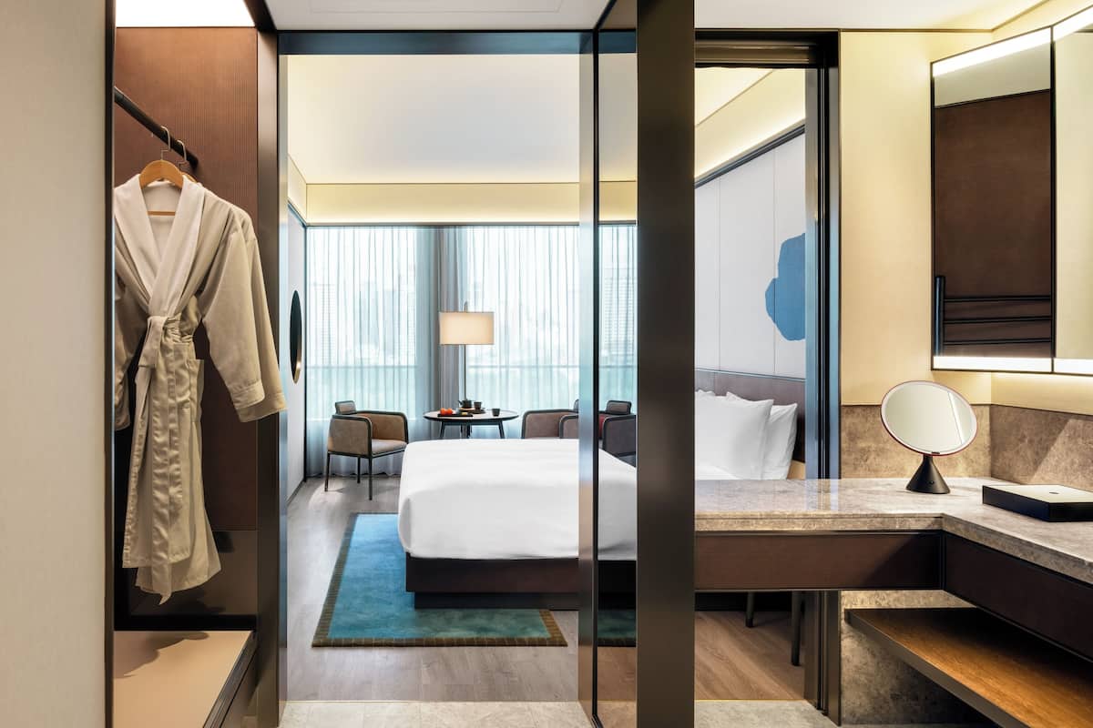 A hotel room with a bed, a desk, a chair, a robe, and a mirror.