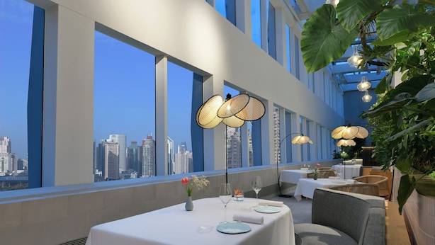 Un comedor moderno con mesas dispuestas para dos, amplias ventanas con vistas a la ciudad y elegantes lámparas colgantes.