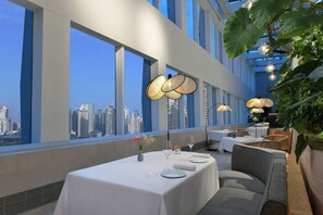 A modern dining area with tables set for two, large windows offering a city view, and hanging pendant lights.