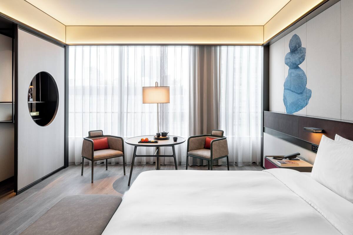 A modern hotel room with a large bed, two armchairs, a small round table, a lamp, and a painting on the wall.