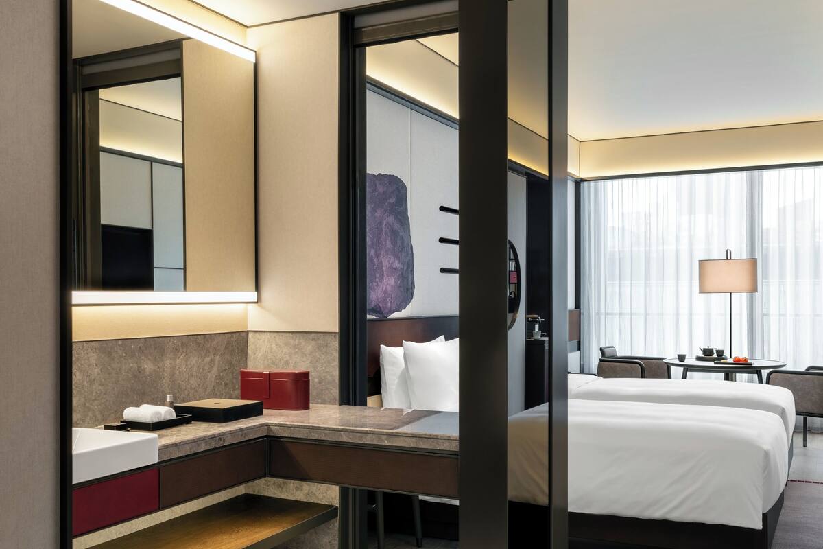 A modern hotel room with a large bed, a desk with a chair, a bathroom with a sink and mirror, and a large window with curtains.