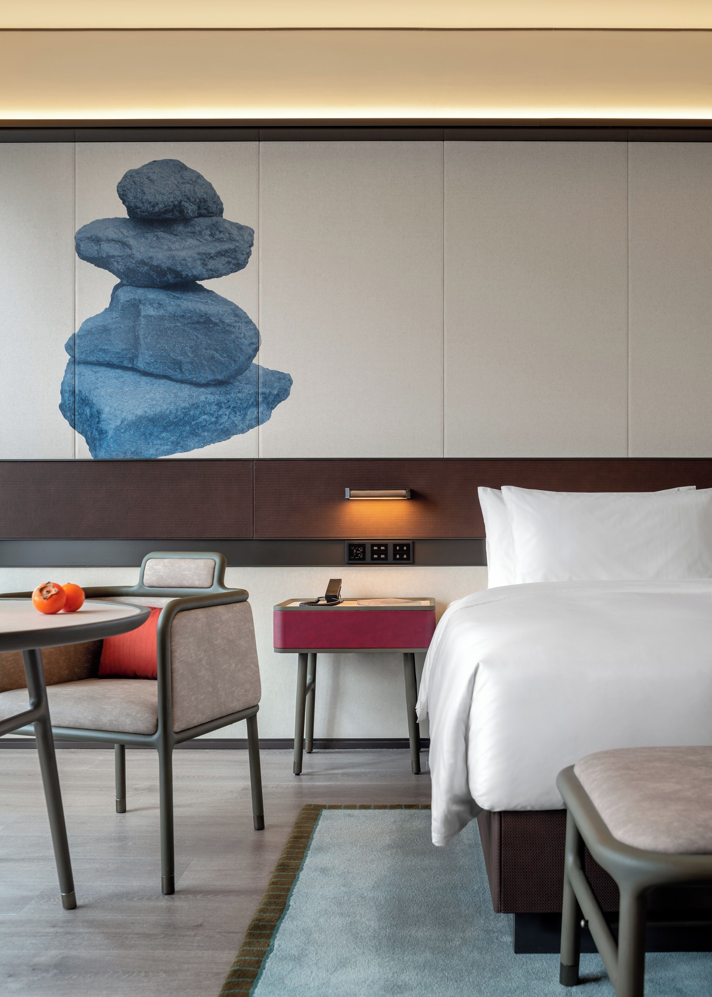 Ein Hotelzimmer mit einem Bett, einem Schreibtisch mit Stuhl, einem kleinen Tisch mit zwei Orangen und einer Wand mit einem blauen, abstrakten Gemälde.