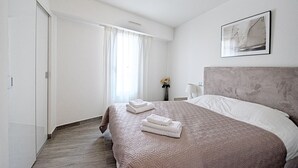 A hotel room with a large bed, a headboard, a bedside table with a lamp, and a window with curtains. - 7 Croisette - Apartment 601 (Cannes)
