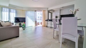 A modern living room with a sofa, a TV, and a glass coffee table. There is a kitchen area with a microwave and cabinets. - 7 Croisette - Apartment 601 (Cannes)