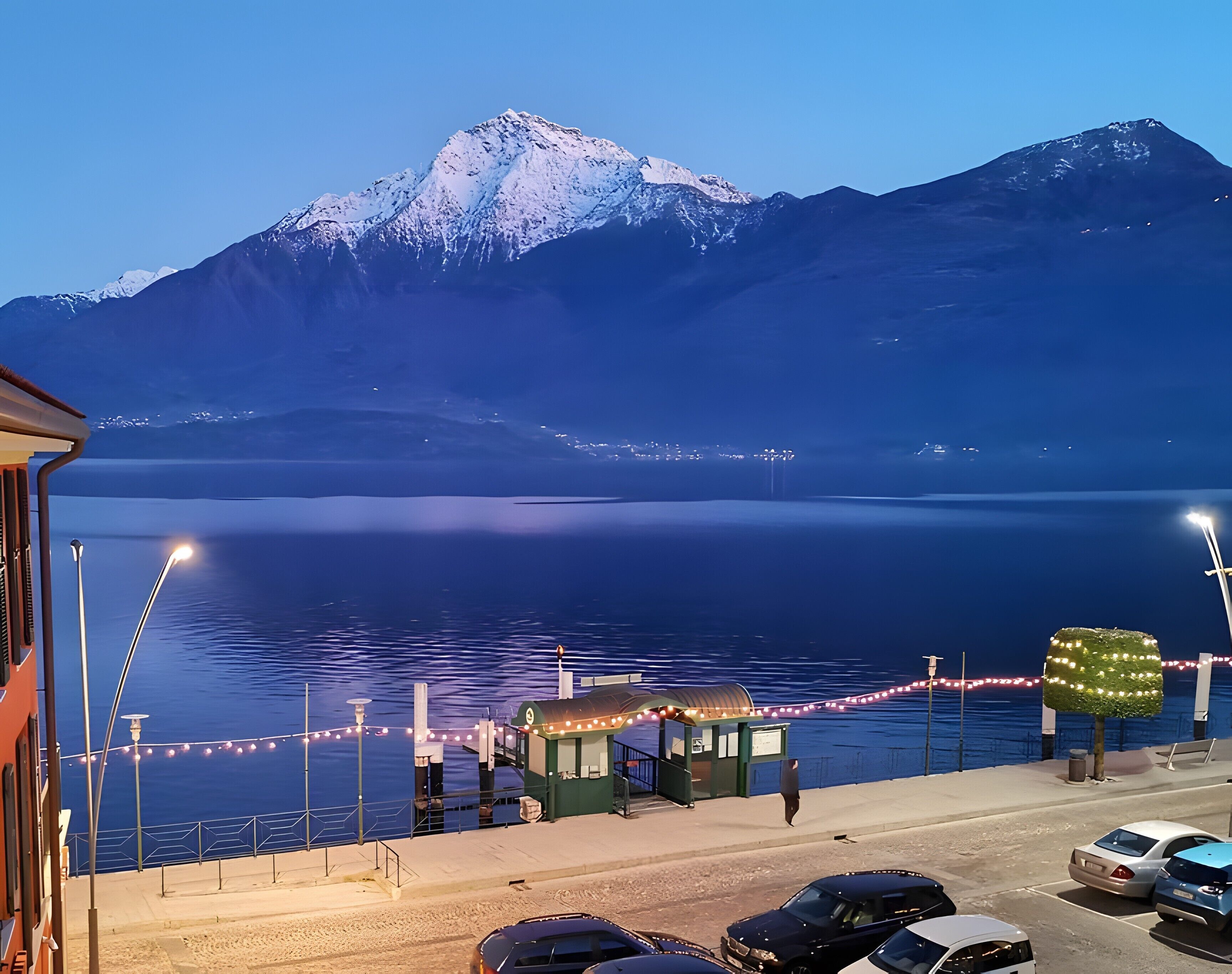 A waterfront view with a mountain in the background, a small kiosk, and parked cars.
