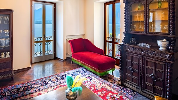 Une pièce avec un fauteuil rouge, un tapis à motifs, un meuble en bois doté de portes en verre et une vue sur l’extérieur, visible à travers une porte.