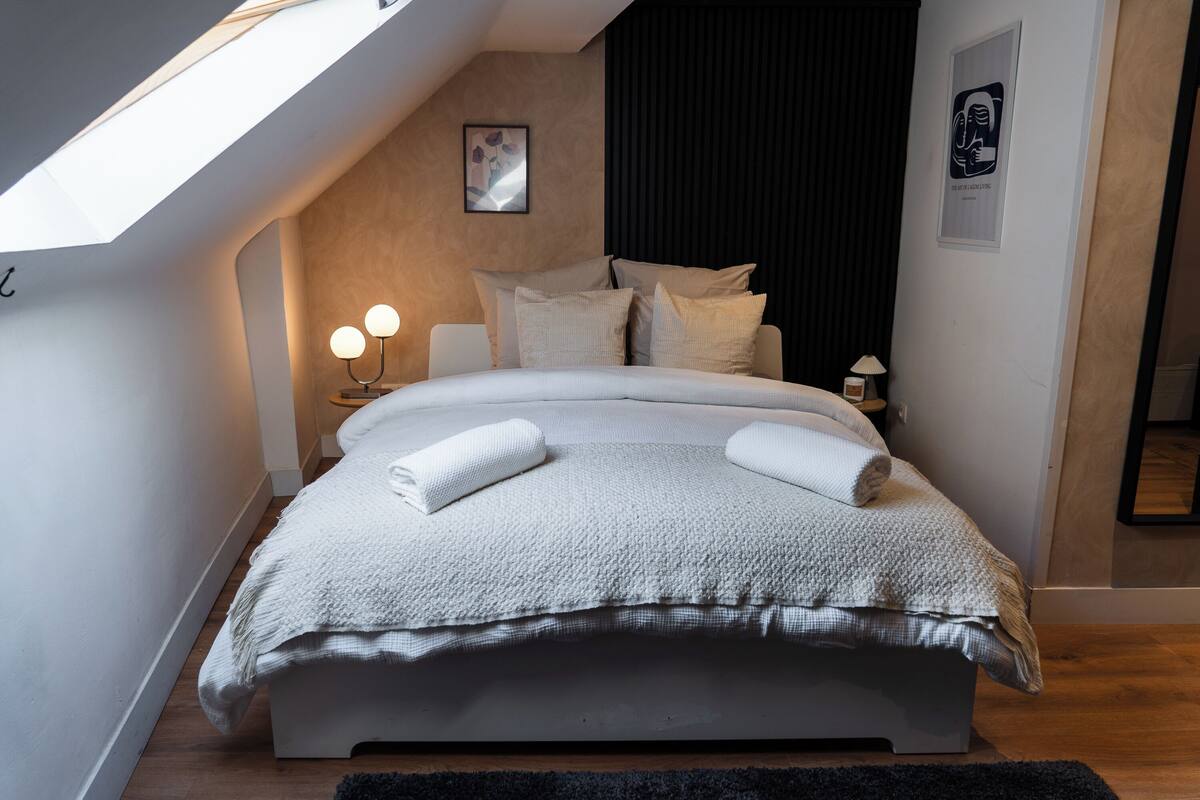 Quarto moderno com claraboia, cama com manta texturizada, duas luminárias de cabeceira e cortina escura.