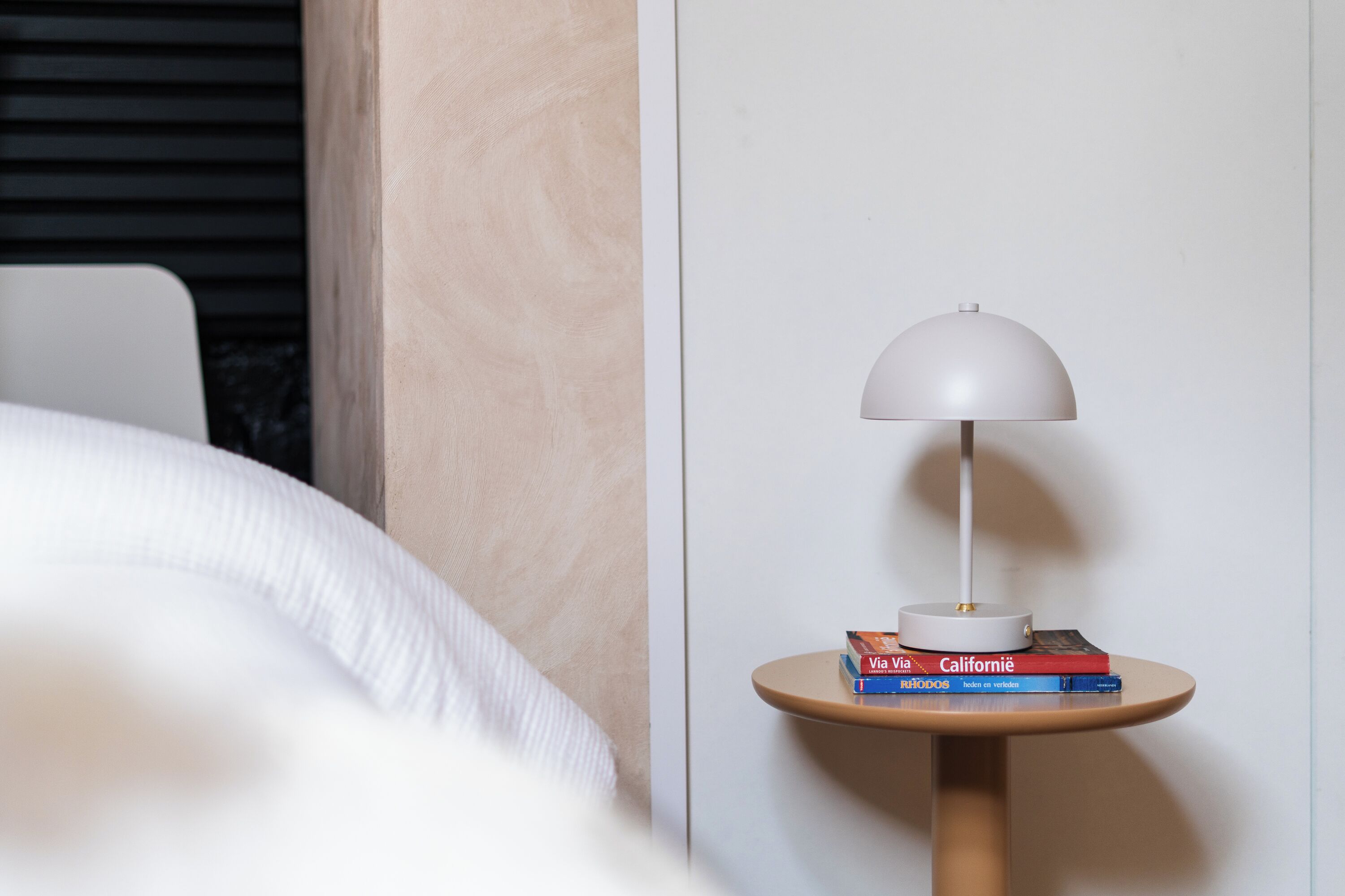 Una cama bien hecha con edredón blanco, una mesita de noche con lámpara y dos libros titulados 'Via Via' y 'California'.