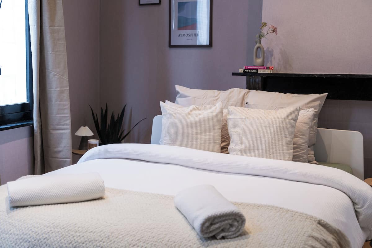 Cama bem arrumada com lençóis e almofadas brancas, mesa de cabeceira com abajur e um quadro na parede.