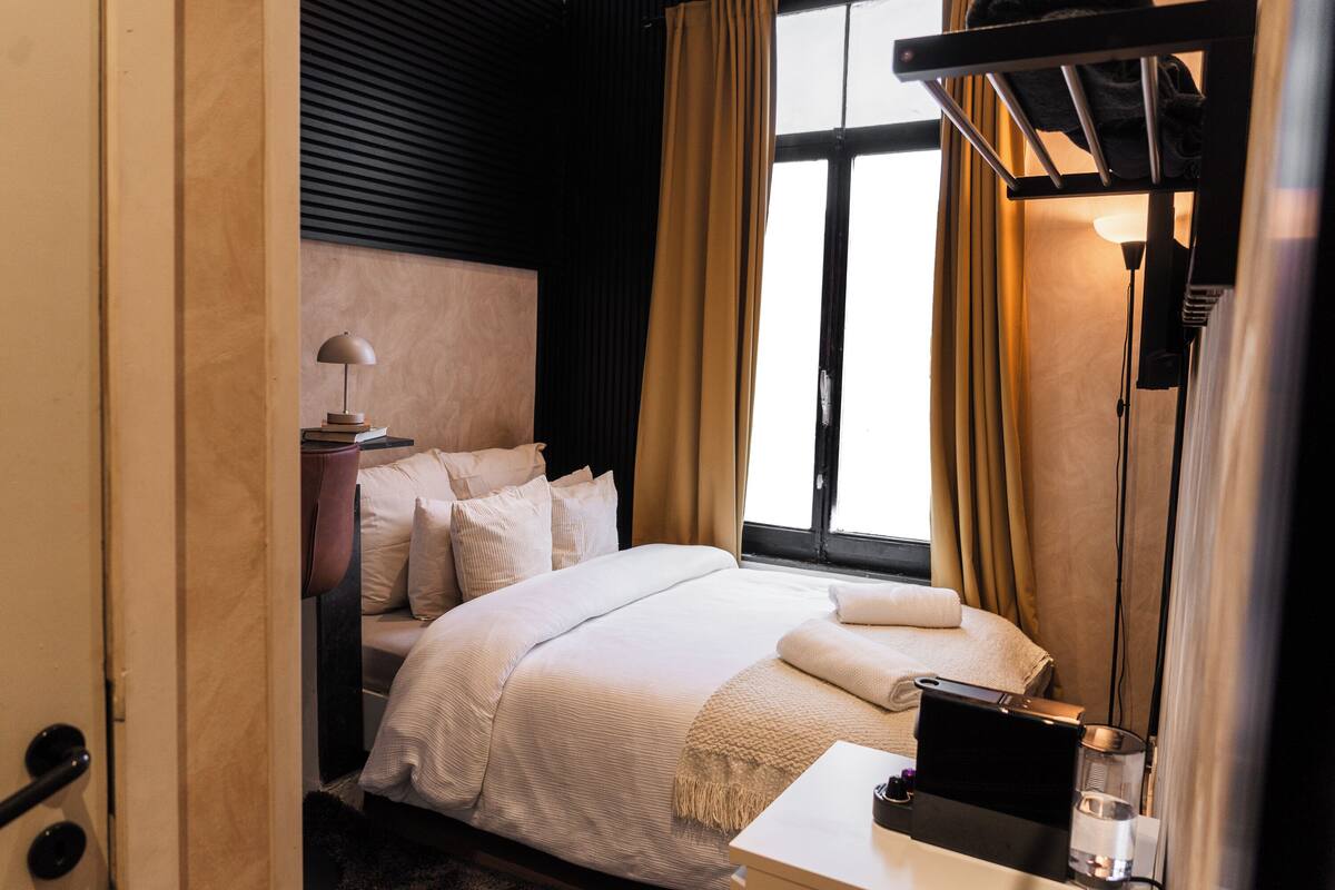 Quarto de hotel com cama, mesa de cabeceira, abajur e janela com cortinas.