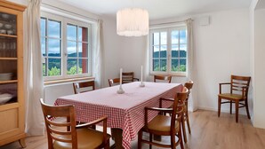 Comedor con una mesa dispuesta para seis, sillas de madera, una araña y ventanas amplias con vistas a la vegetación.