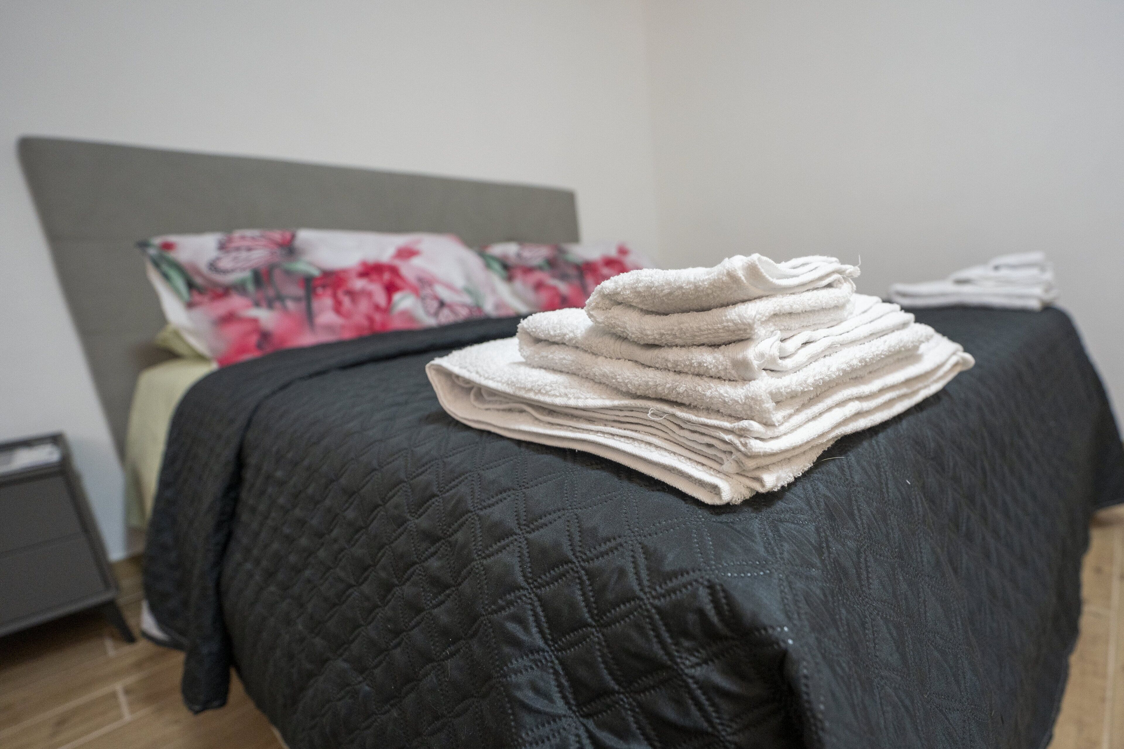 A neatly made bed with a dark quilted bedspread and a stack of folded towels on the bed.