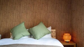 A bed with white bedding and two green pillows, a wooden nightstand with a lit orange lamp, and a bamboo-patterned wall.