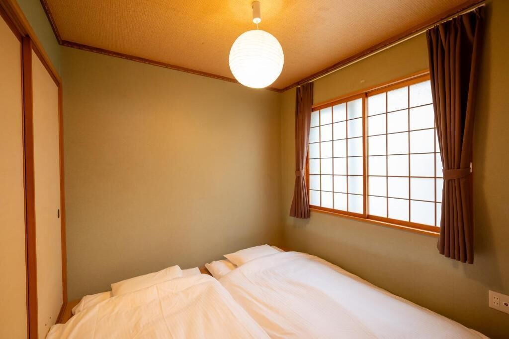 A small, single-bed room with a wooden ceiling, a window with curtains, and a hanging light fixture.