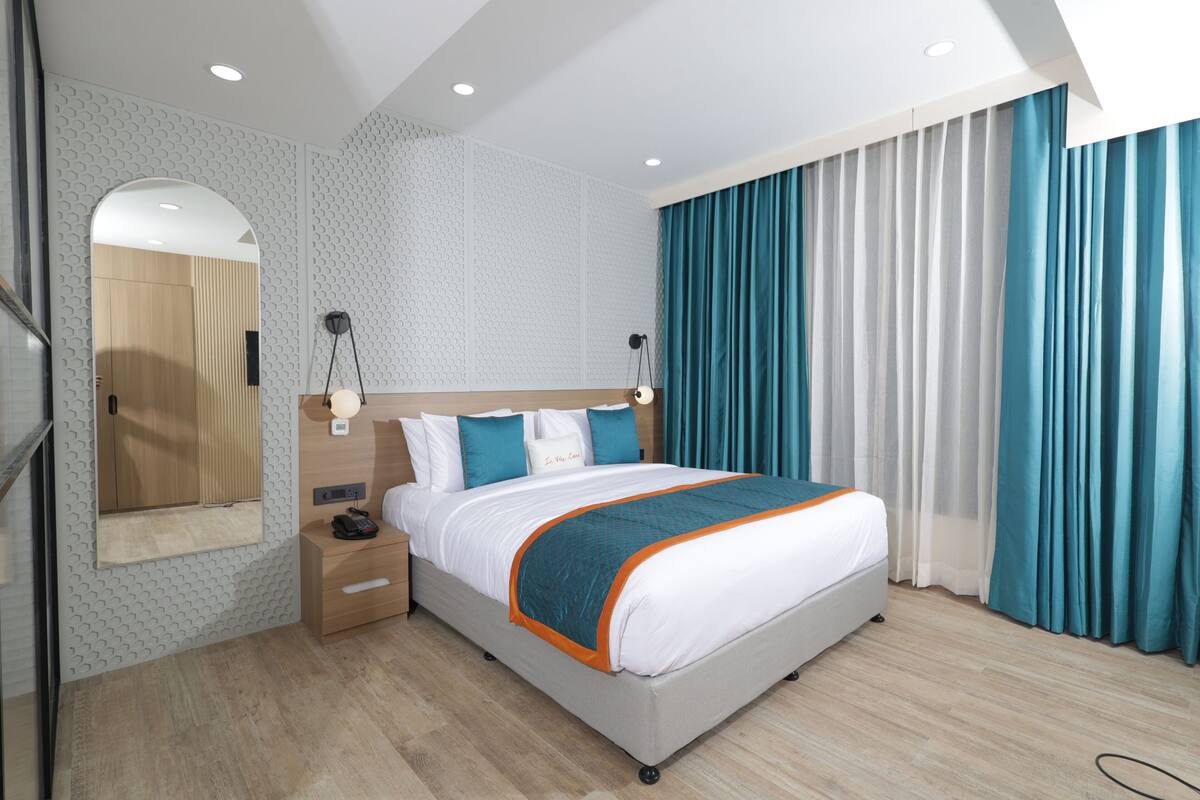A modern hotel room with a large bed, a bedside table, a nightstand, a mirror, and a window with curtains.