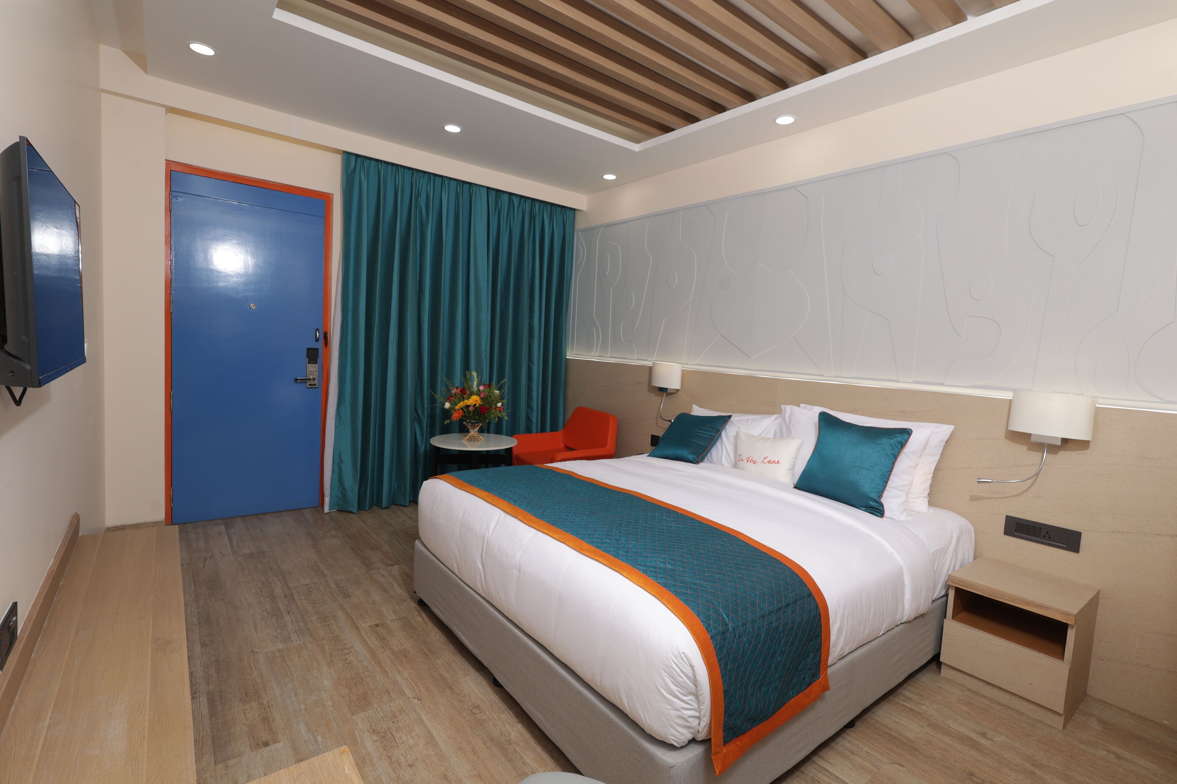 A modern hotel room with a large bed, a desk, a chair, a TV, and a door.
