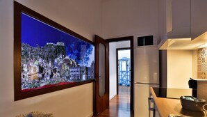 A hallway with a large window displaying a cityscape, a kitchen area with a refrigerator, and a dining table with a placemat. - GuestHost - La Casa del Presepe Central  House (Naples)