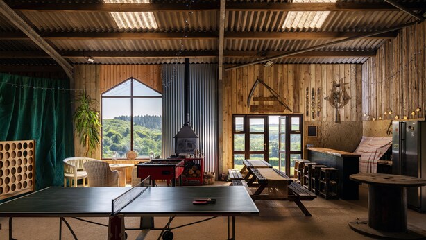 A rustic wooden room with a ping pong table, a large window with a view of greenery, a hanging light fixture, and a wooden ceiling. - Callestock Courtyard Cottages - Wheal Busy charming detached cottage perfect for your honeymoon (Truro)