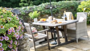 An outdoor patio with a round table and wicker chairs, surrounded by greenery and a stone wall.