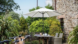 Une terrasse avec une table et des chaises, protégée par un grand parasol, entourée de verdure et bordée d’un mur en pierre.