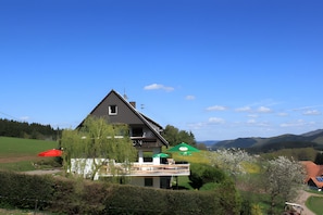 A two-story house with a balcony, surrounded by greenery and a clear sky.