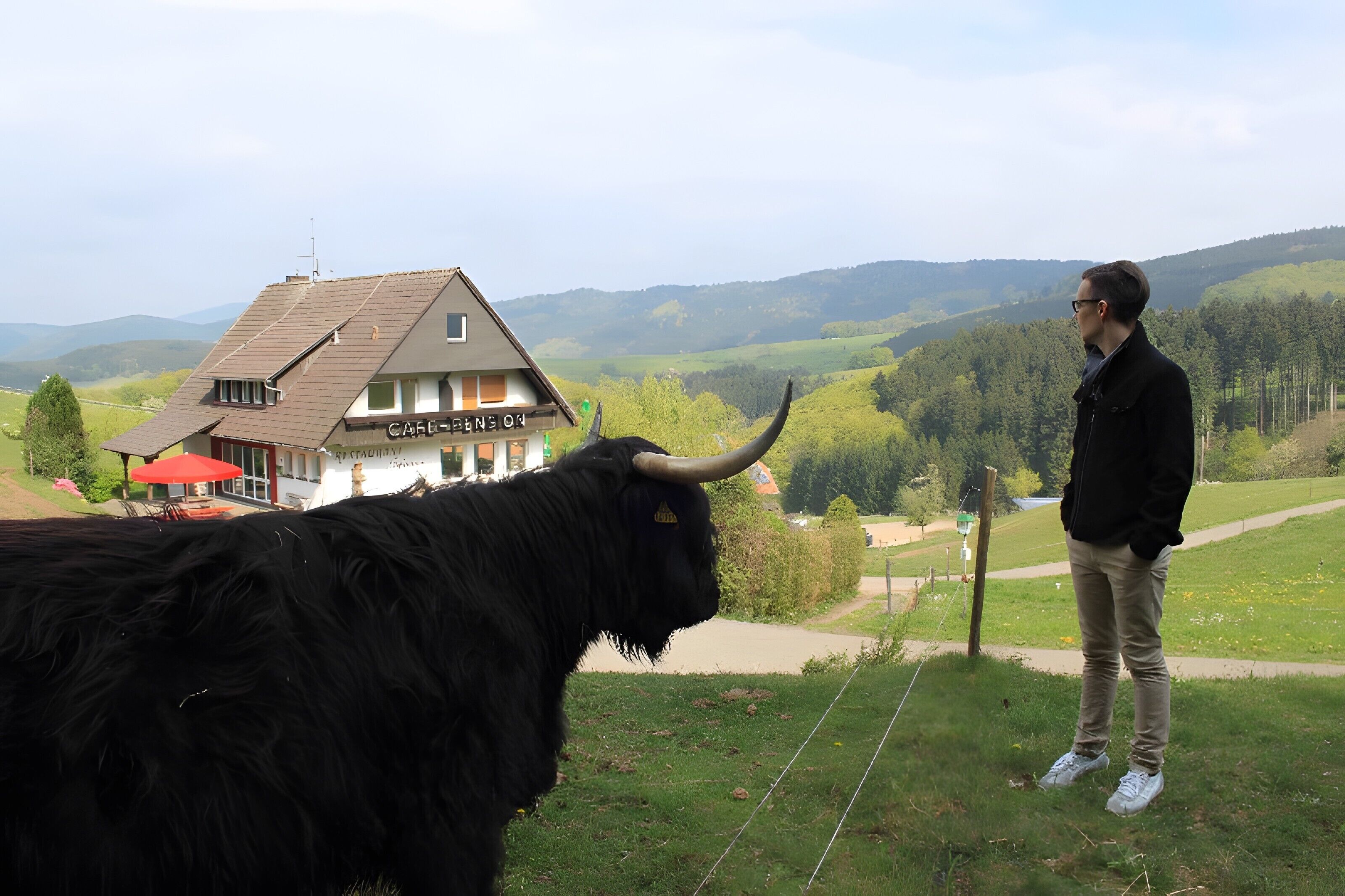 Une personne se tient à côté d’un yak à la longue fourrure et aux grandes cornes, avec un bâtiment en arrière-plan.
