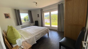 A bedroom with a bed, a chair, a table with a lamp, a window with curtains, and a view of greenery outside. - Room 'No. 4 with Private Balcony' with Shared Terrace, Shared Garden and Wi-Fi (Mühlenbach)