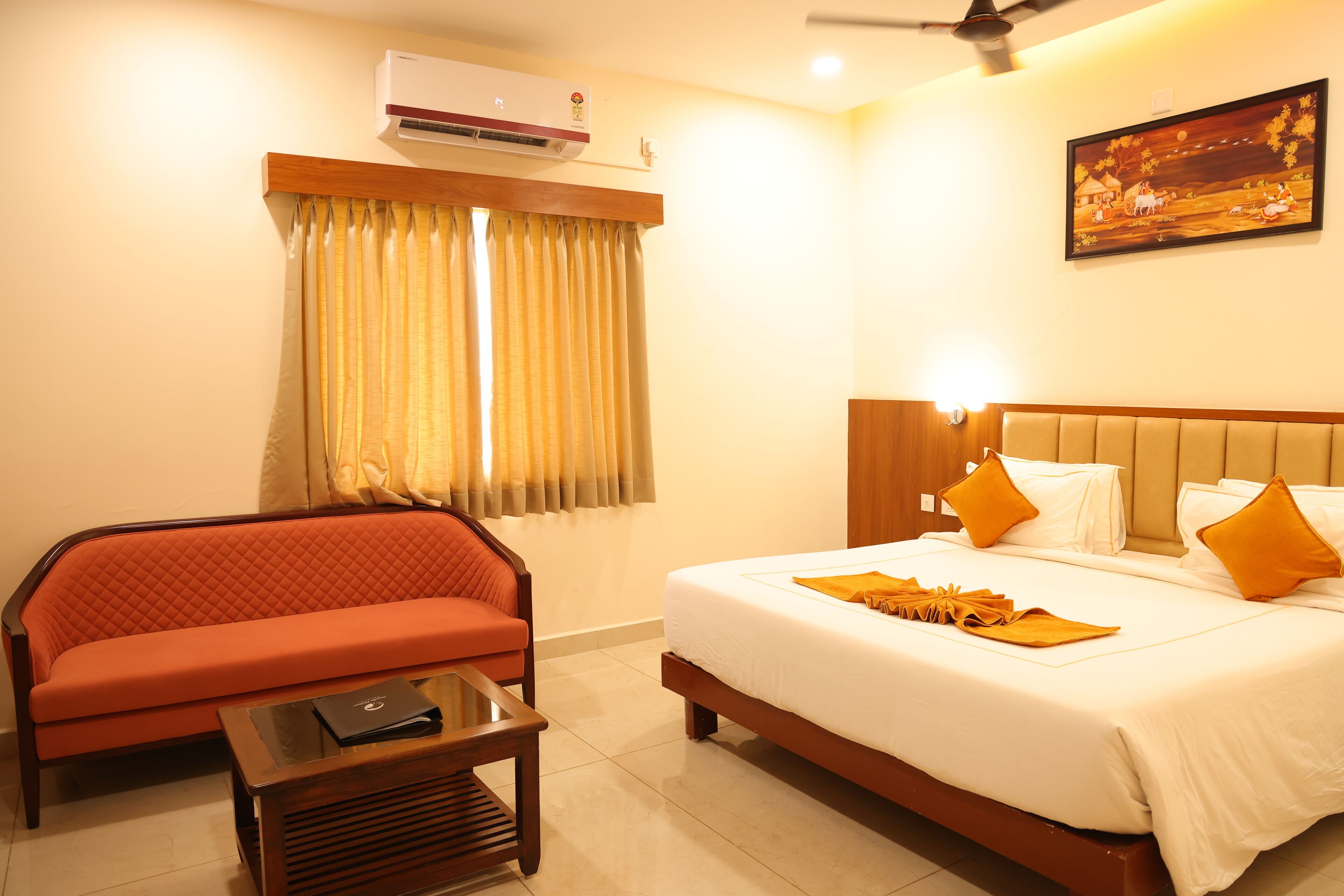 A hotel room with a bed, a sofa, a table, an air conditioner, a painting on the wall, and a window with curtains.