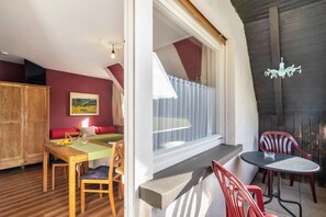 Une pièce comprenant une table à manger en bois, un canapé rouge, une fenêtre avec des rideaux et un petit balcon avec une table et des chaises.