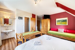 Une chambre d’hôtel avec un lit, une table en bois sur laquelle se trouvent un vase et des fruits, un canapé avec des coussins rouges, une télévision et un tableau accroché au mur.