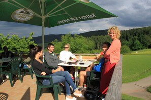 En grupp människor sitter vid ett utomhusbord under ett grönt parasoll, omgivna av gröna stolar, i en miljö med utsikt över en gräsklädd äng och träd.