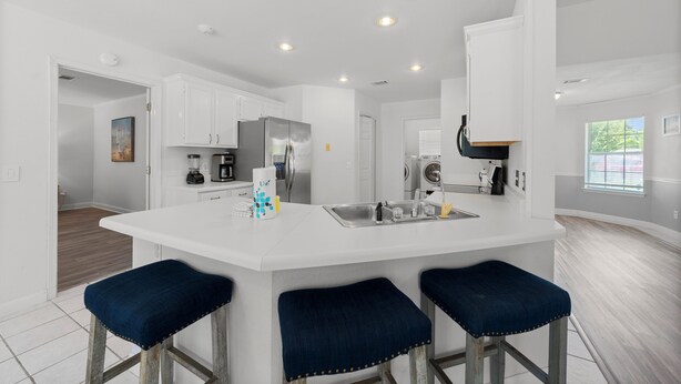 A modern kitchen with white cabinetry, a stainless steel refrigerator, a built-in oven, and a large island with bar stools. - Tranquil Haven | Pet Friendly (Pensacola)
