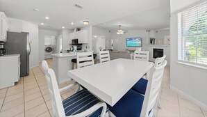 A modern kitchen with white cabinetry, a dining table with blue cushioned chairs, and a television in the background. - Tranquil Haven | Pet Friendly (Pensacola)