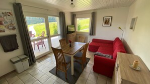 A room with a dining table, chairs, a red sofa, a TV, and a window with curtains. - Room 'No. 2 with Private Balcony', Shared Garden and Wi-Fi (Mühlenbach)