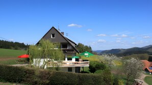 A two-story house with a balcony, surrounded by greenery and a clear sky. - Room 'No. 2 with Private Balcony', Shared Garden and Wi-Fi (Mühlenbach)