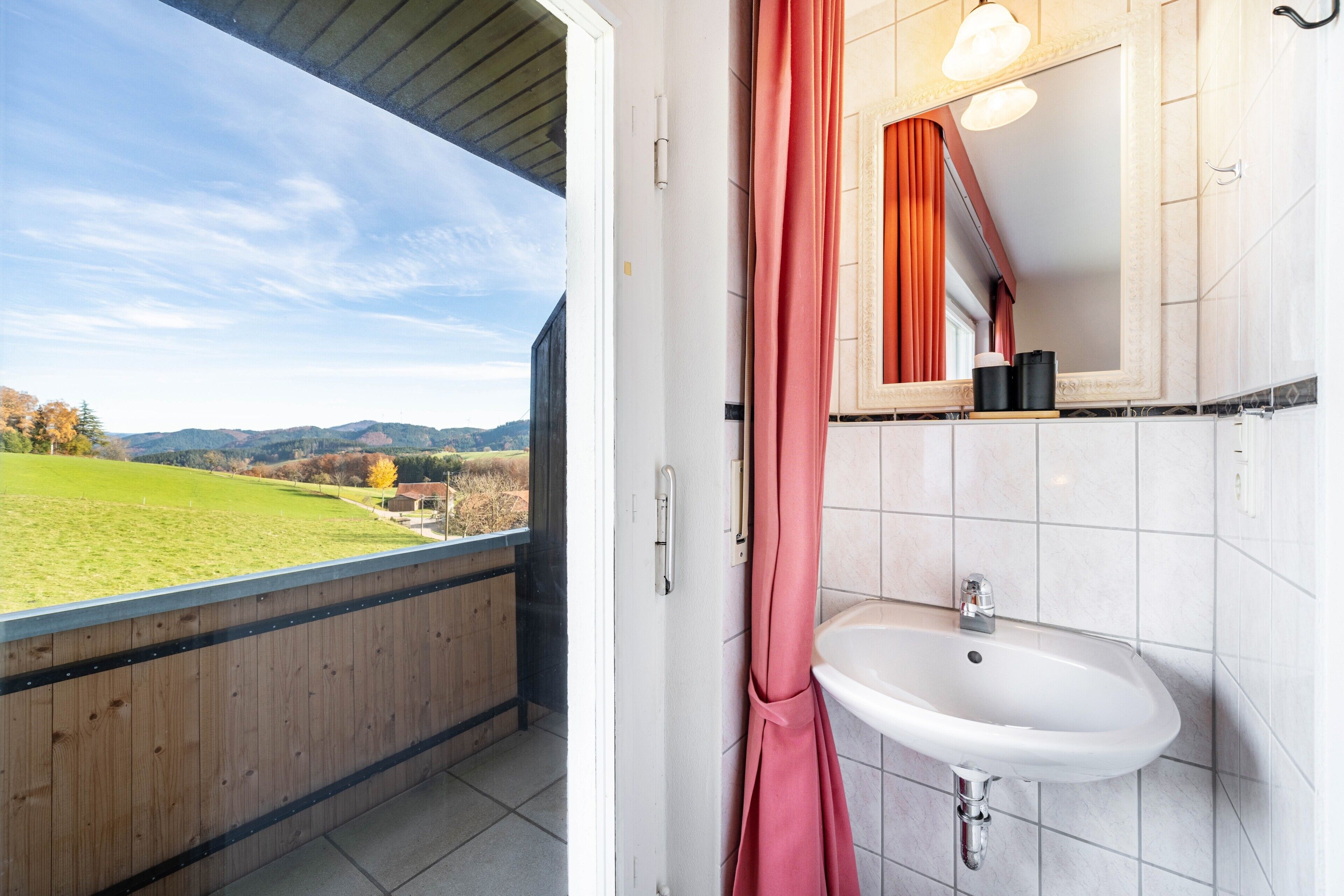 A bathroom with a white sink, a mirror, and a view of a green field and mountains.